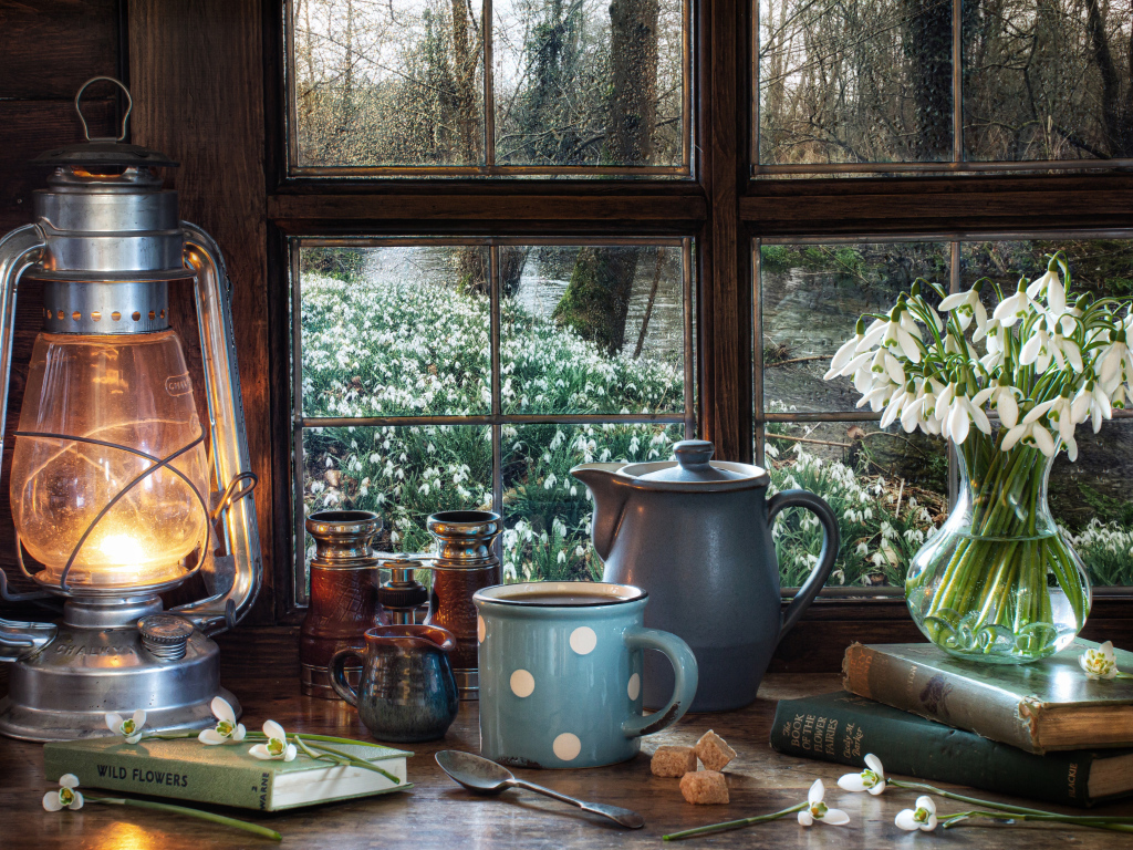 Kerosene lamp on the table with a mug, books and a bouquet of snowdrops by the window