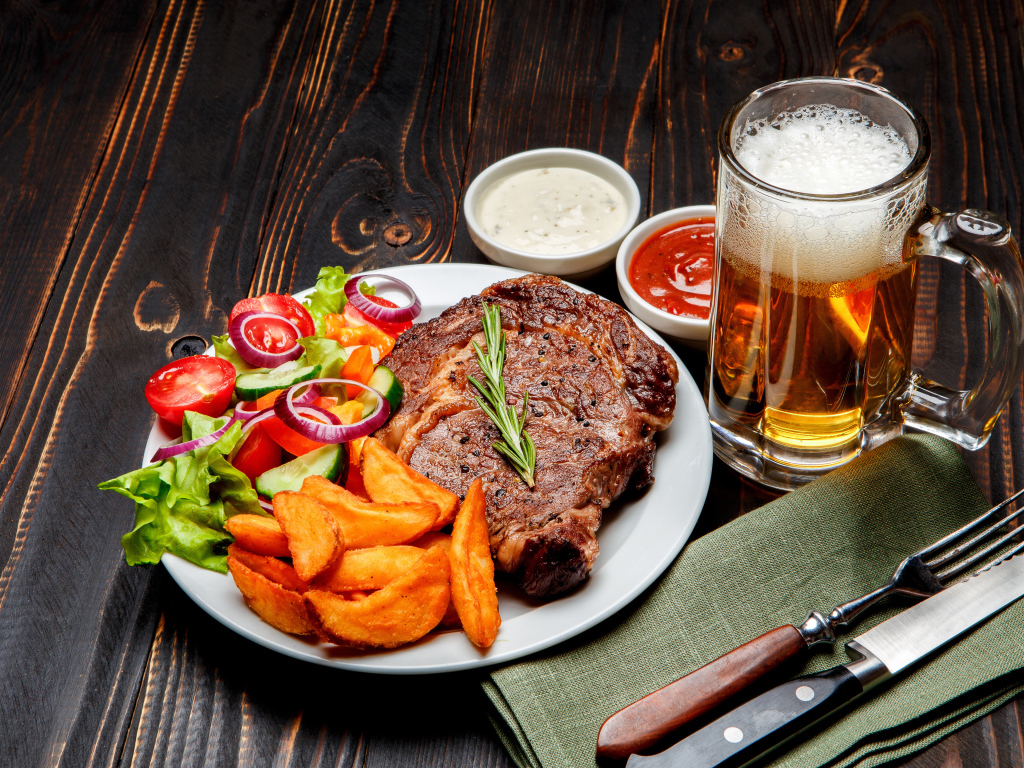 Кусок мяса на тарелке с картофелем и салатом на столе с соусом и пивом