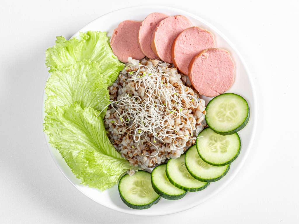 Гречка на тарелке с колбасой, огурцами и листьями салата