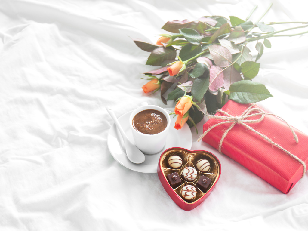 Чашка кофе на белой кровати с конфетами, подарком и букетом роз
