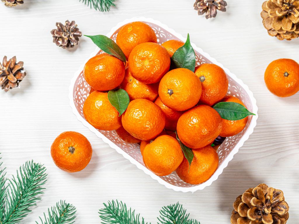 Много спелых мандаринов в белой корзинке на столе с шишками