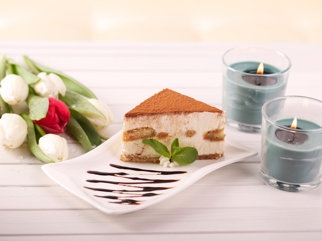Кусок тирамису на столе с зажженными свечами и букетом тюльпанов