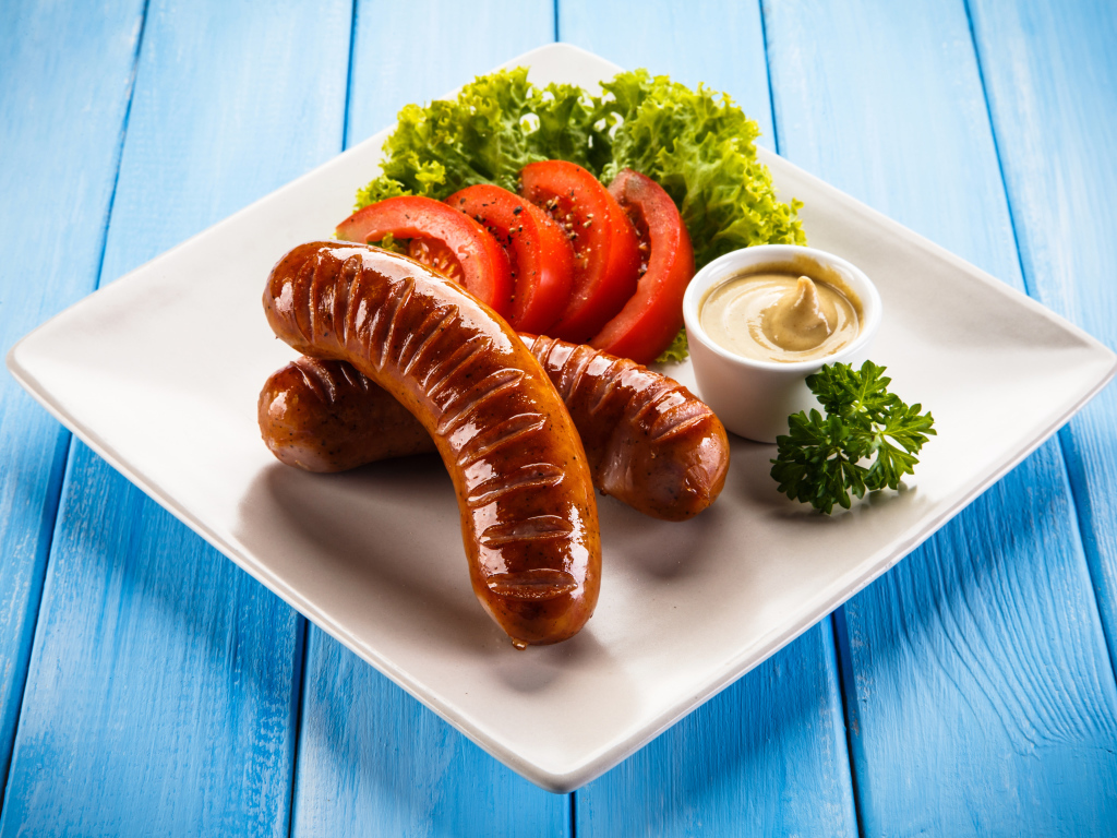 Две жареные колбаски на тарелке с помидорами, горчицей и листьями салата 