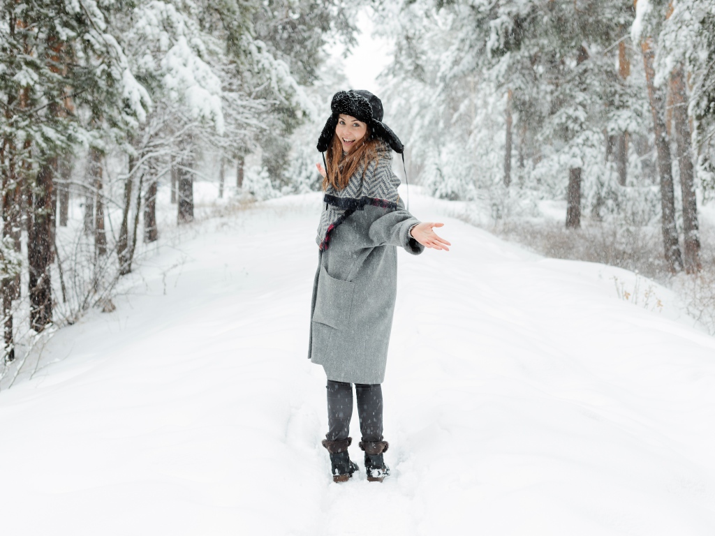 Девушка в пальто и теплой шапке гуляет в заснеженном лесу 