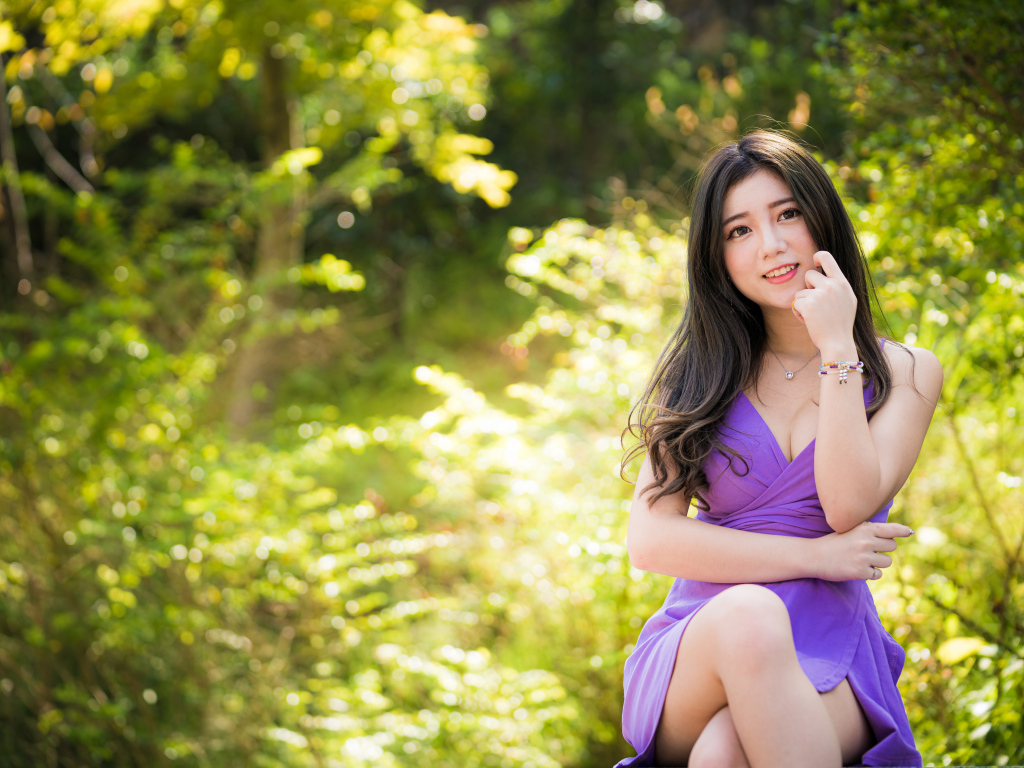 Улыбающаяся девушка азиатка в фиолетовом платье сидит в парке 
