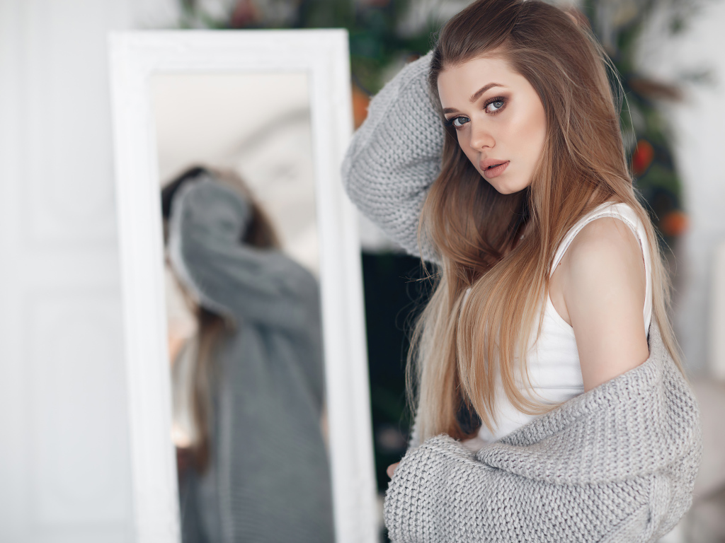 Молодая девушка в серой кофте стоит у зеркала 