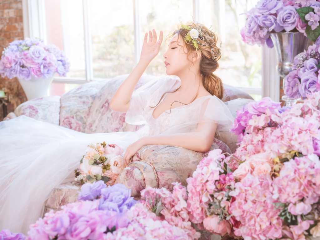 Девушка азиатка в белом платье сидит на диване в комнате с цветами