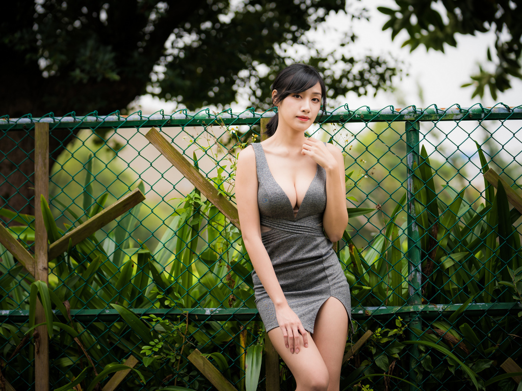 Красивая азиатка в сером платье стоит у зеленой сетки 