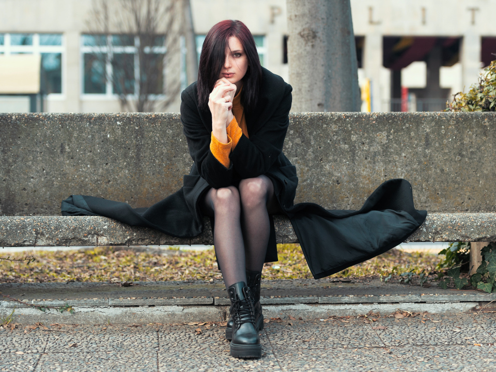 Девушка в черном пальто сидит на бетонной лавке 