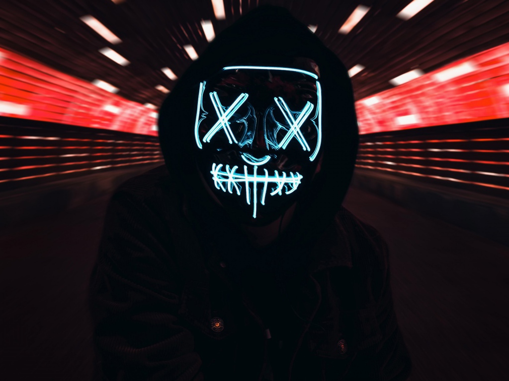 Парень в черной куртке в неоновой маске анонимуса на лице