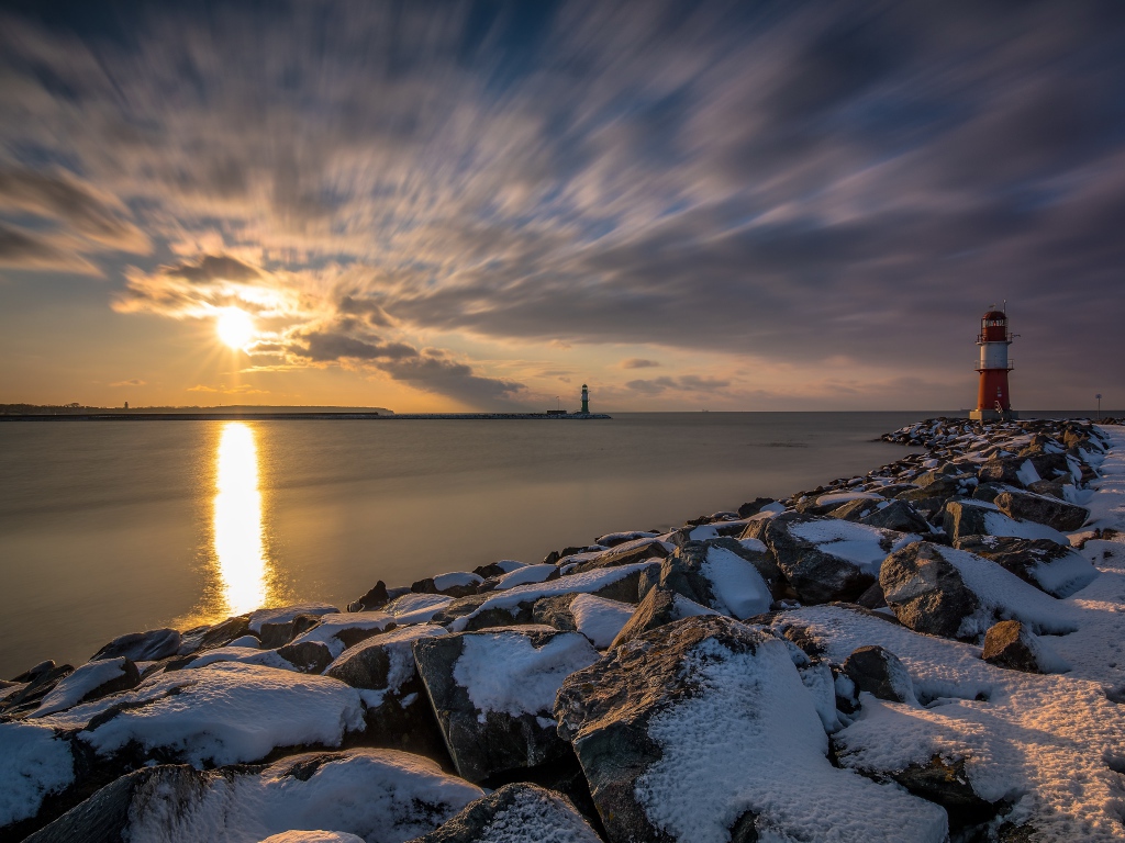 Заснеженные камни на берегу моря с маяком на закате