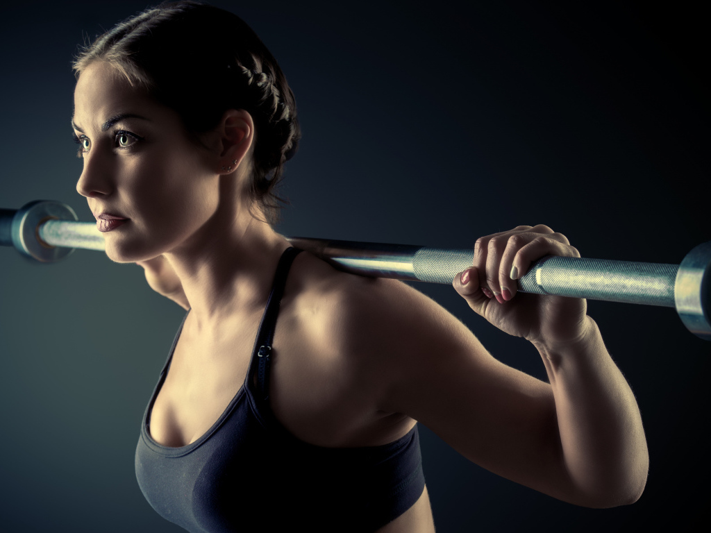 Спортивная девушка занимается со штангой в спортзале на сером фоне