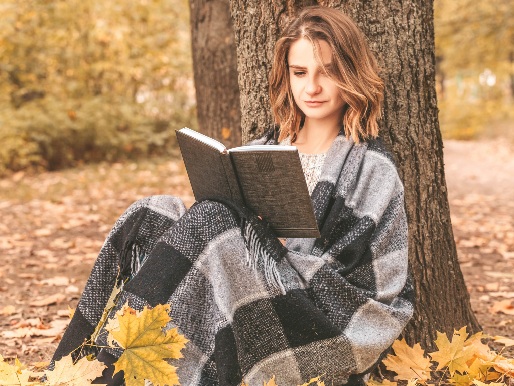 Девушка в пледе сидит с книгой под деревом