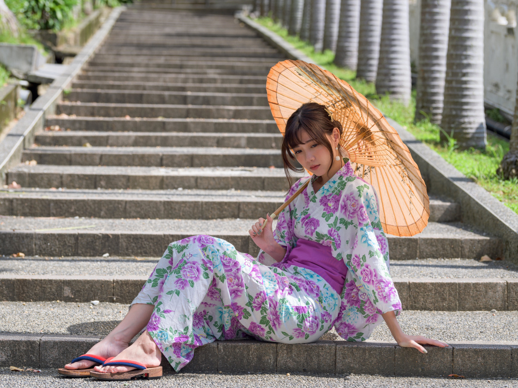 Девушка азиатка в кимоно с зонтом в руках сидит на ступеньках 