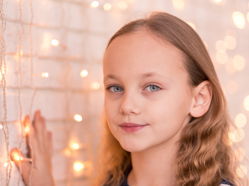 Красивая голубоглазая девочка стоит у стены с гирляндой