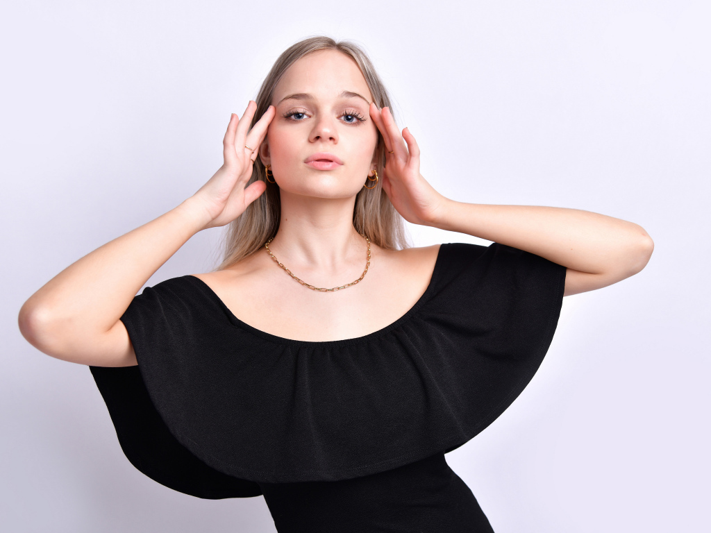 Девушка в черном платье с руками у головы на белом фоне