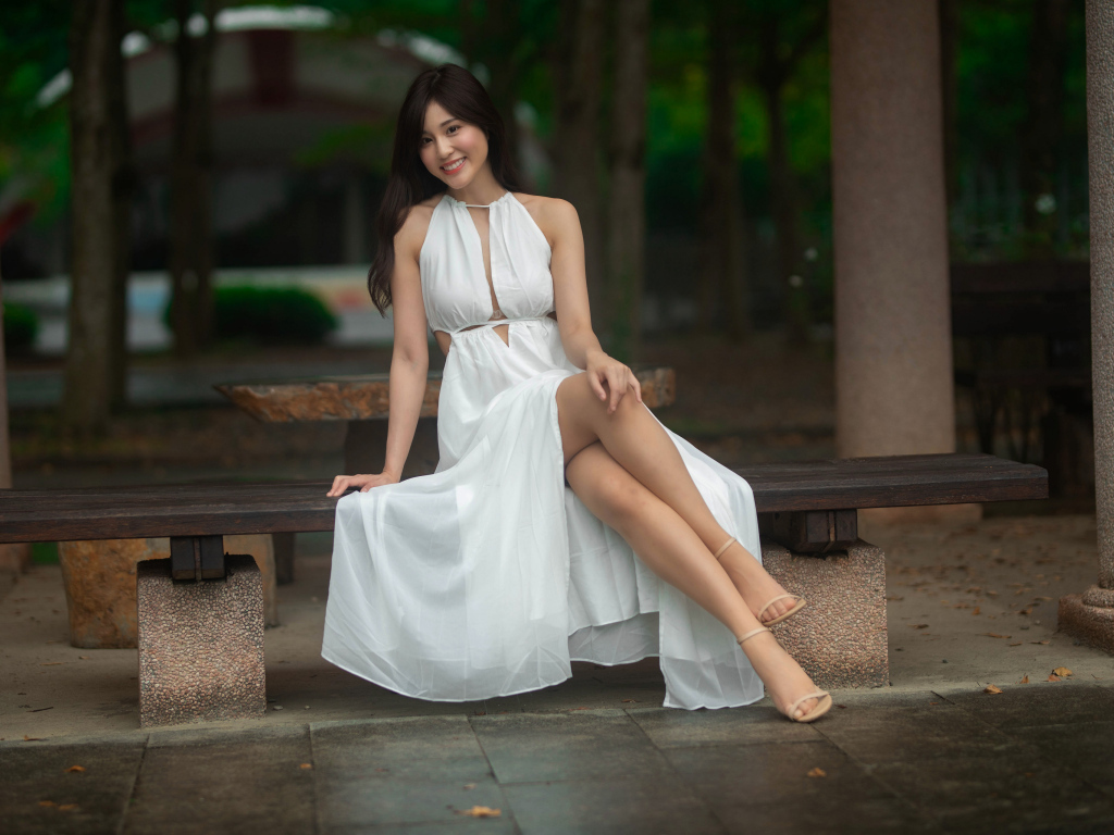 Улыбающаяся азиатка в белом платье сидит на лавке