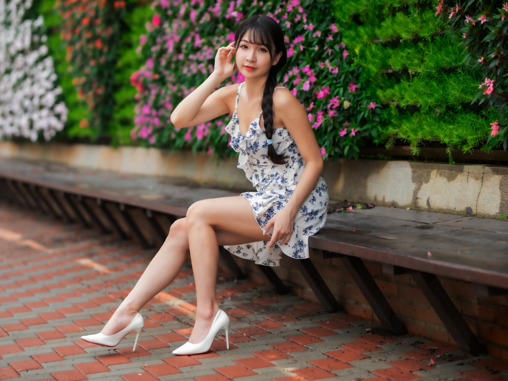 Азиатка в платье сидит на лавке у цветущих растений