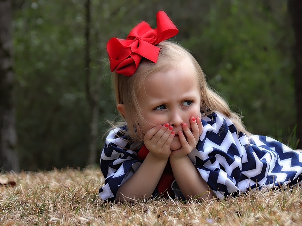 Маленькая девочка с красным бантом на голове лежит на траве