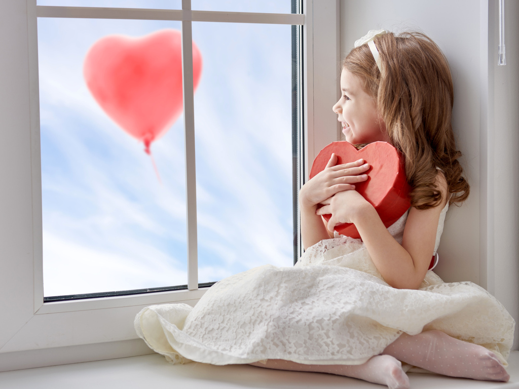 Маленькая девочка с коробкой в форме сердца сидит у окна