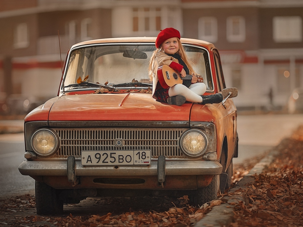Маленькая девочка с гитарой сидит на красном автомобиле осенью
