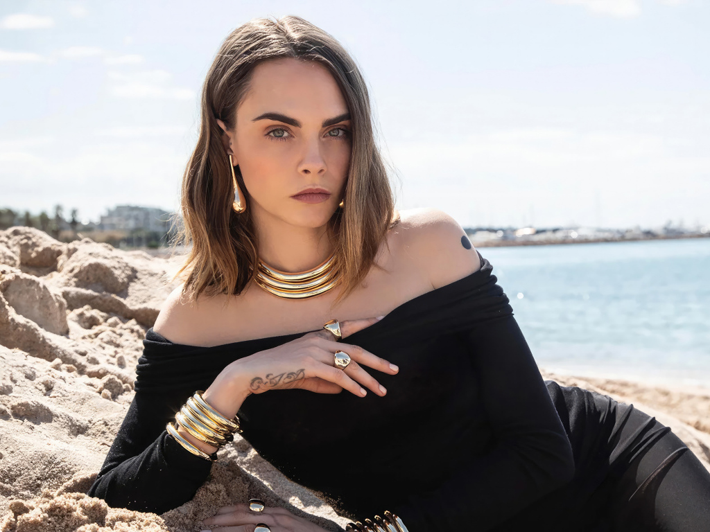 Кара Делевинь в черном платье лежит на песке