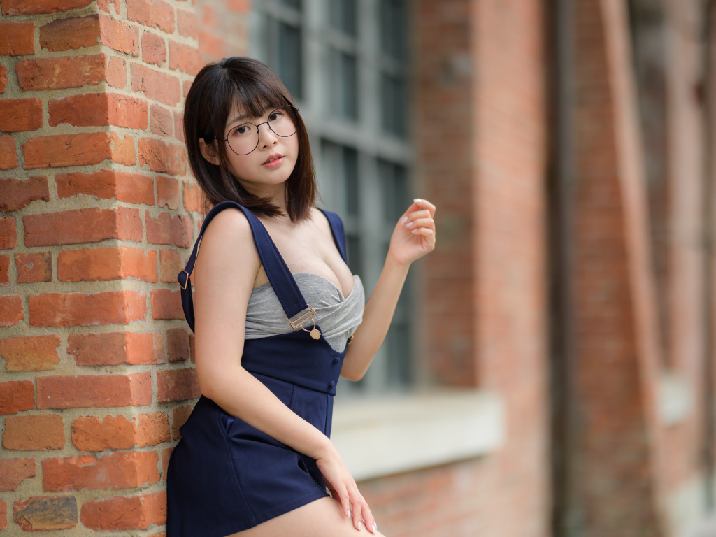 Азиатка в очках стоит у кирпичной стены