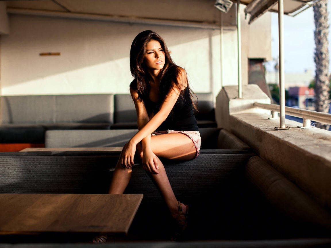 Брюнетка сидит на спинке дивана и смотрит в окно