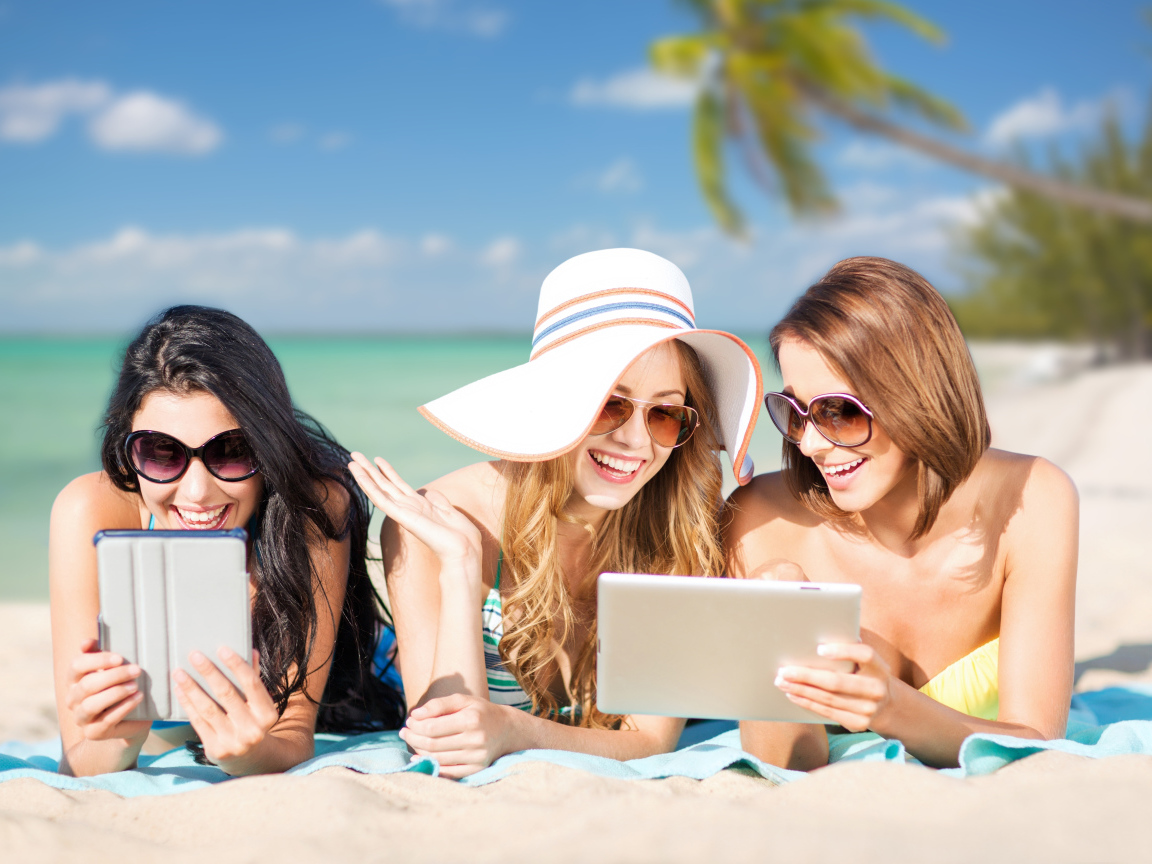 Три улыбающиеся девушки загорают на пляже с планшетами в руках