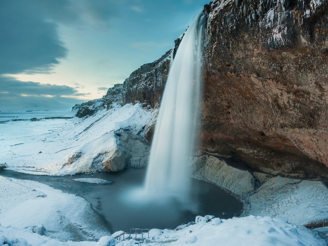 Водопад стекает с утеса на фоне заснеженной природы, Исландия
