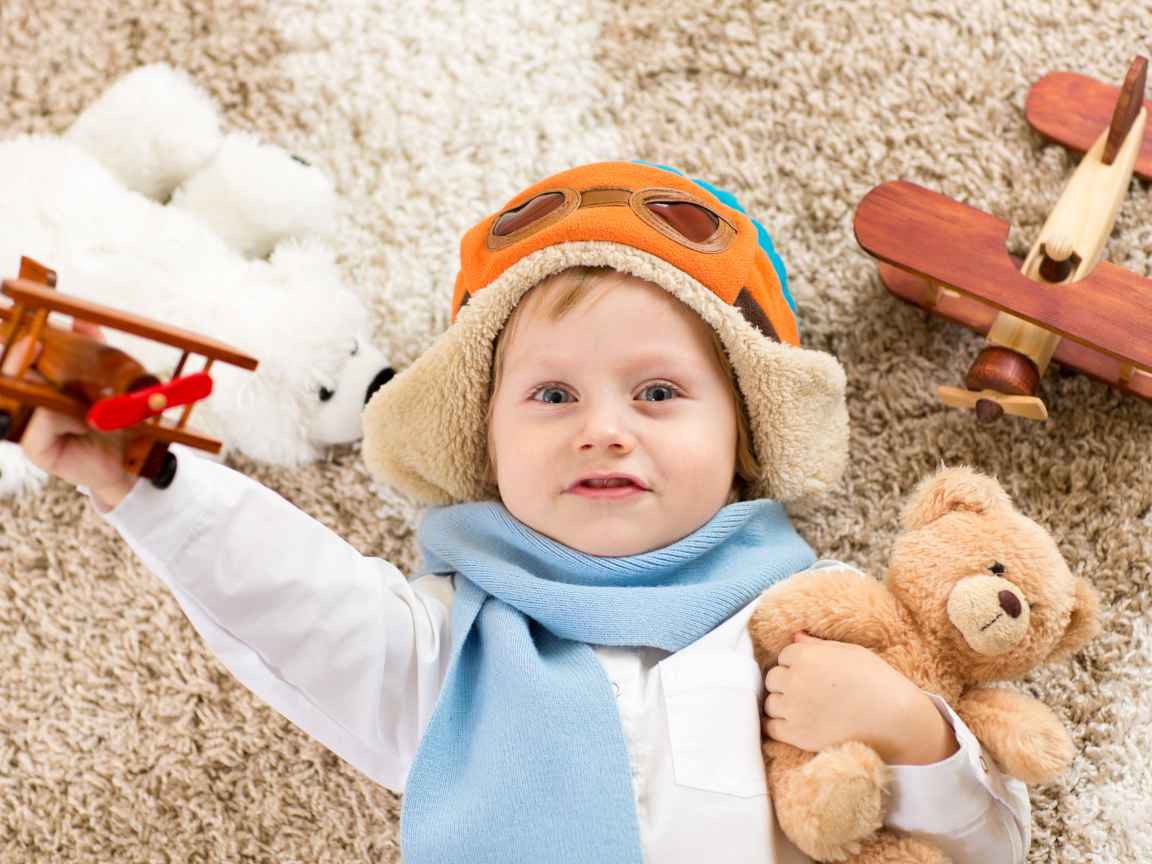 Маленький мальчик в шапке и игрушечным самолетом и медвежонком в руках