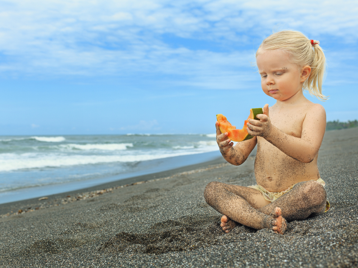 Маленькая девочка сидит на песке у моря с кусочком дыни