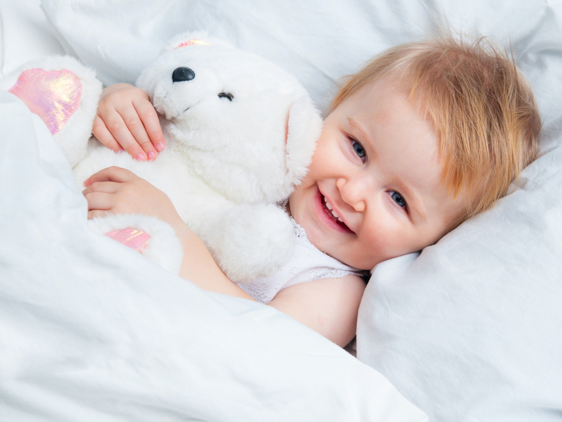 Улыбающийся ребенок лежит в кровати с белым плюшевым медвежонком