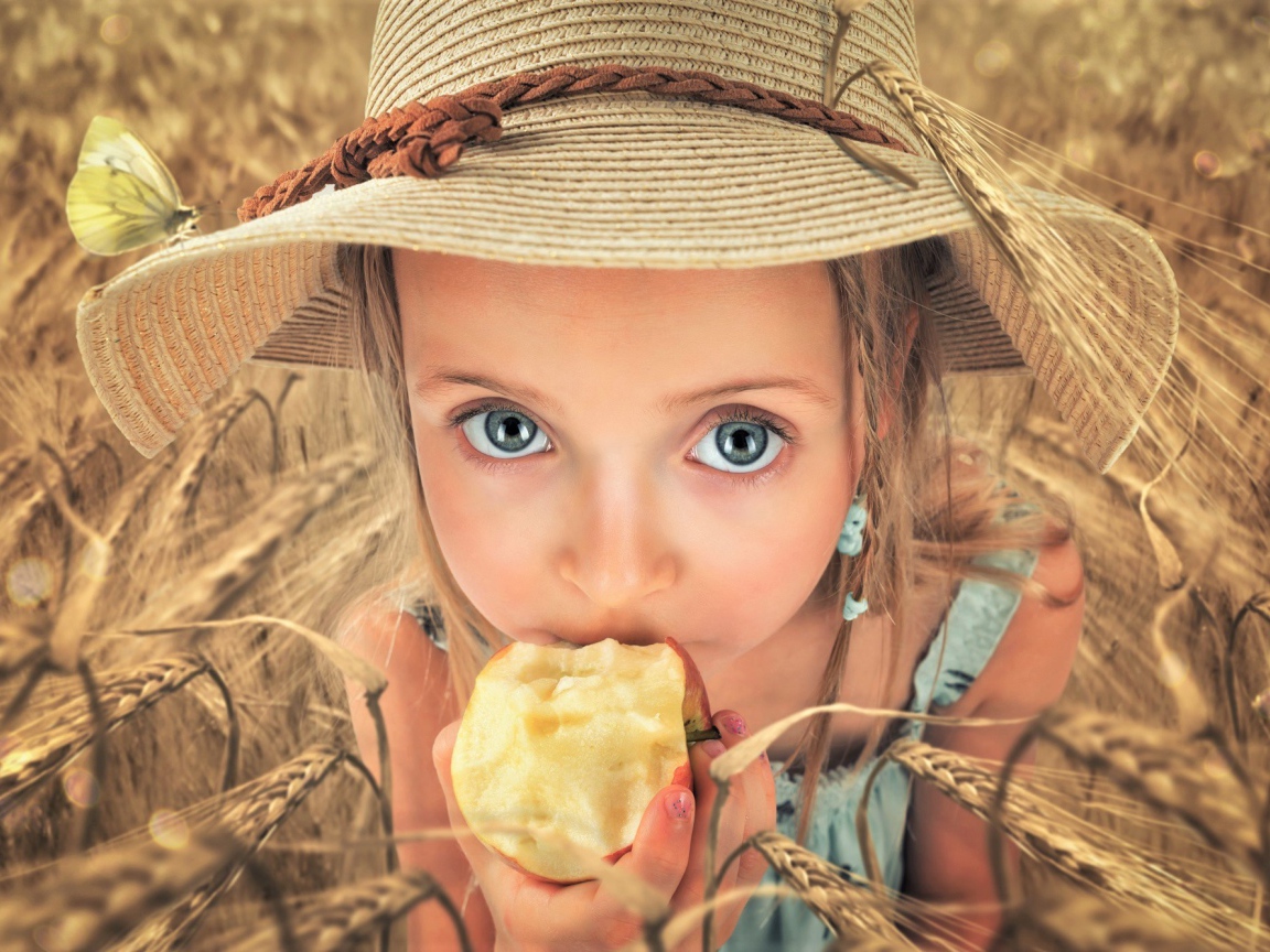 Маленькая голубоглазая девочка в шляпе ест яблоко