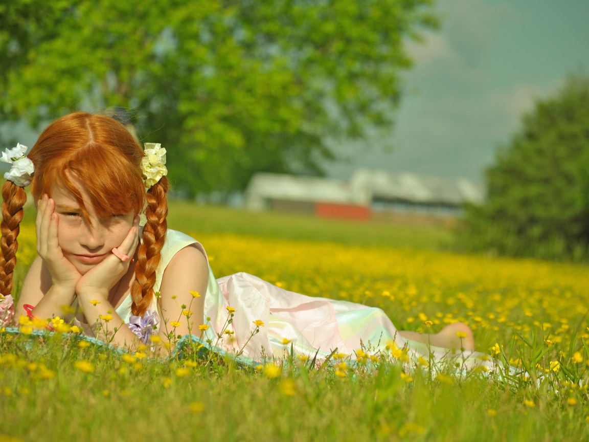 Рыжеволосая девочка с косичками лежит на зеленой траве