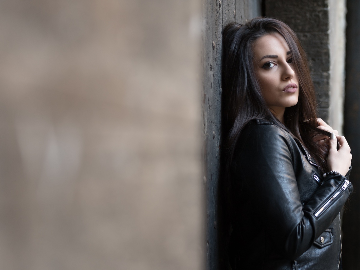 Красивая  брюнетка в кожаной куртке стоит у стены