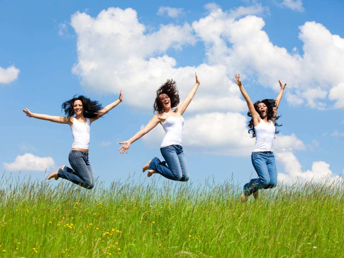 Три молодые девушки прыгают в зеленой траве на фоне неба 
