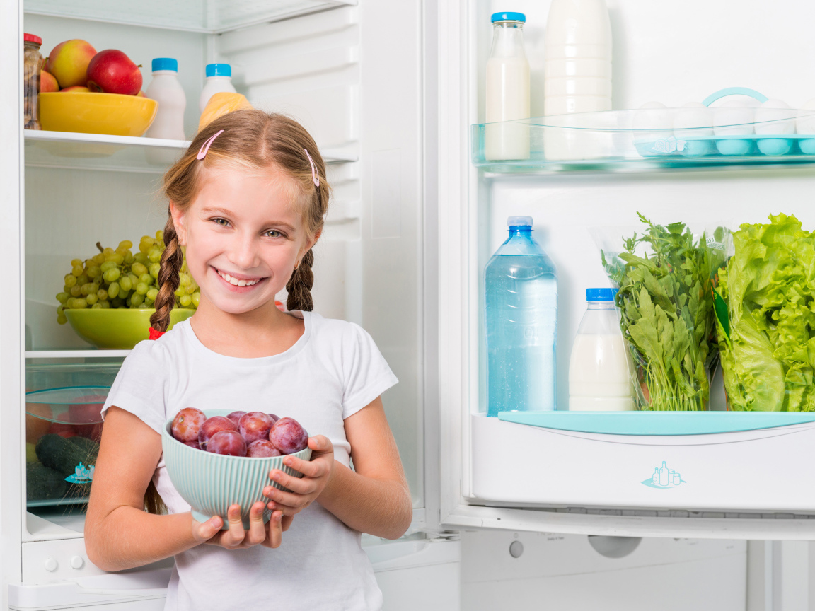 Маленькая девочка с миской со сливами в руках у холодильника 