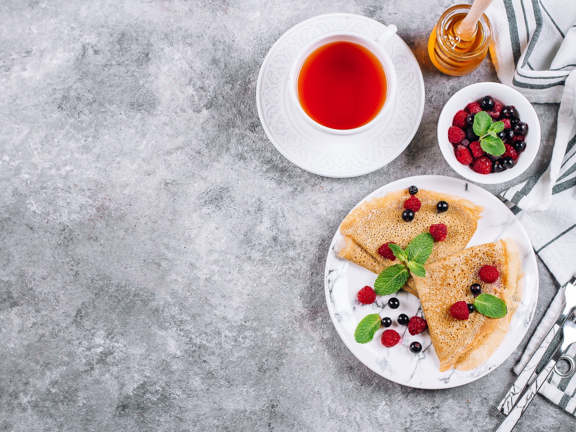 Тонкие блинчики на столе с ягодами, чаем и медом