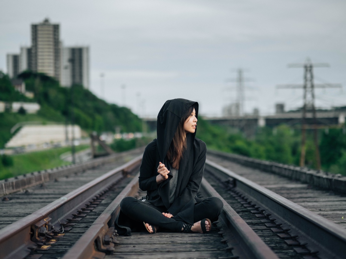 Девушка азиатка в черном балахоне сидит на рельсах 