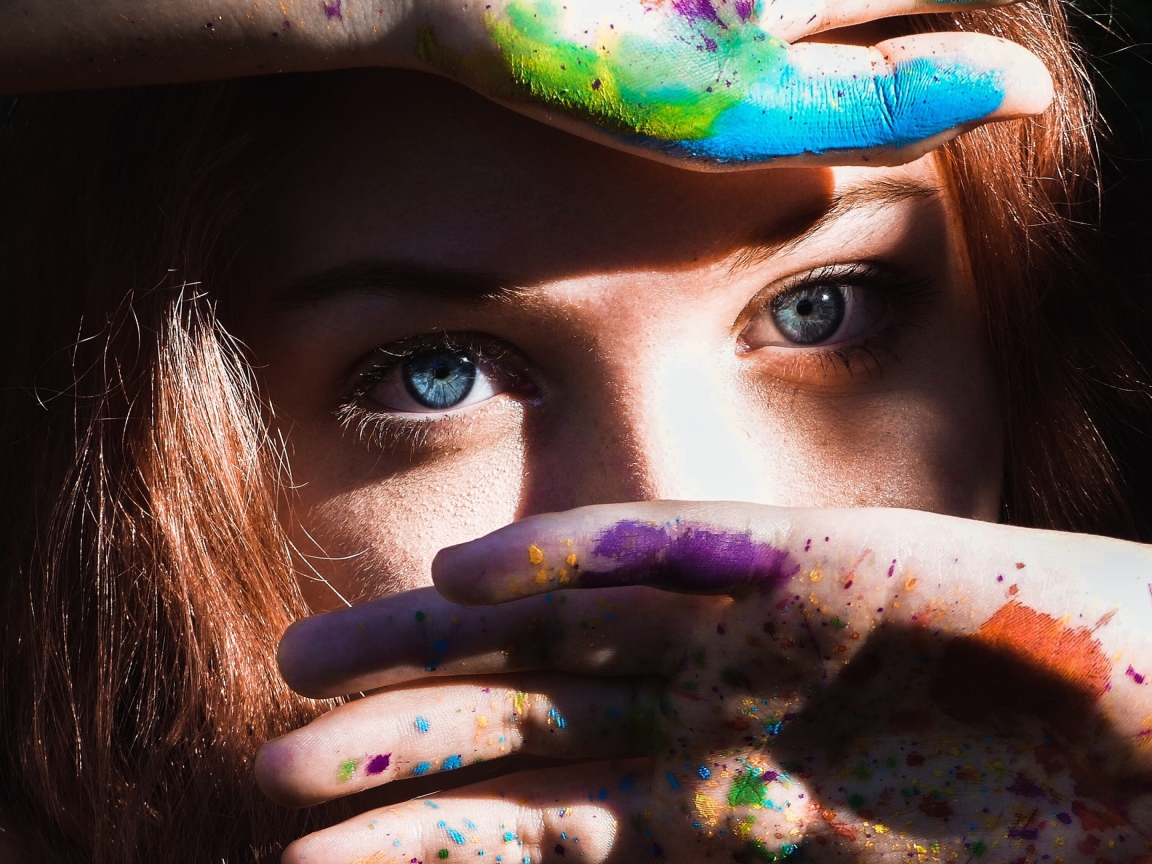 Красивая голубоглазая девушка с руками в разноцветной краске у лица