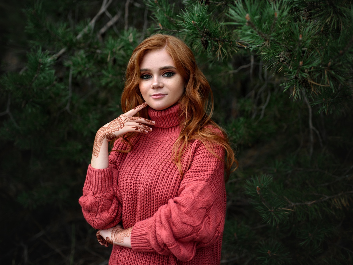 Рыжеволосая девушка в розовом свитере стоит у ели