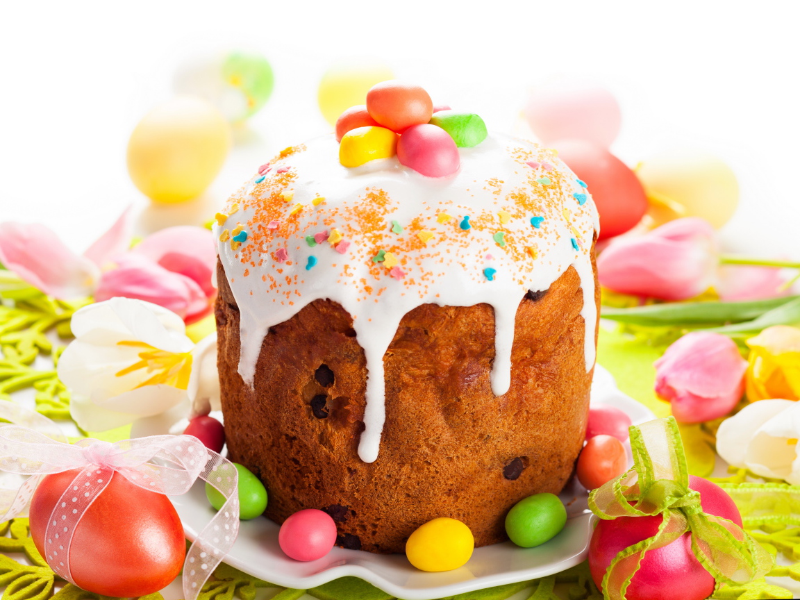 Красивый пасхальный кулич с белой глазурью и конфетами на столе с яйцами