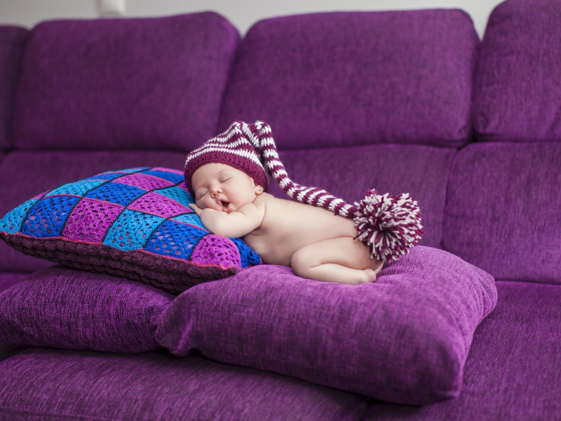 Маленький ребенок в большой шапке спит на фиолетовых подушках