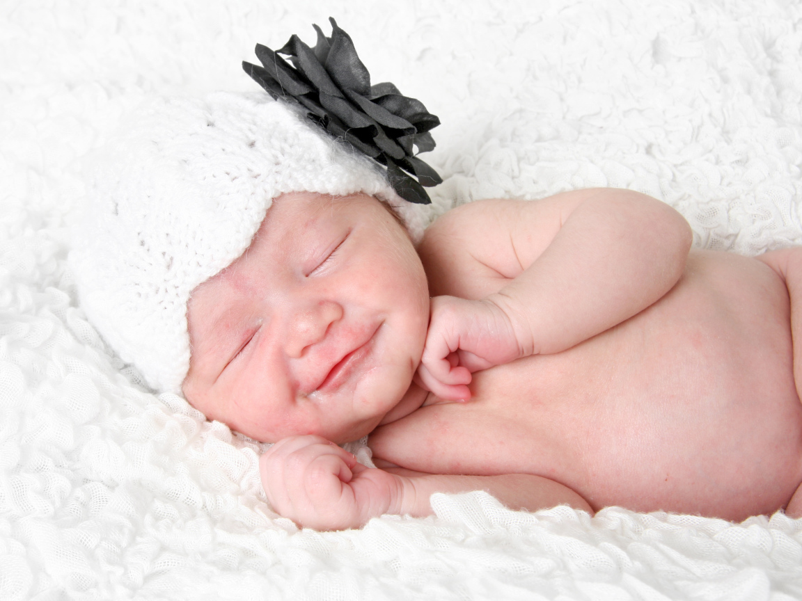 Улыбающийся спящий ребенок в белой вязаной шапке с цветком