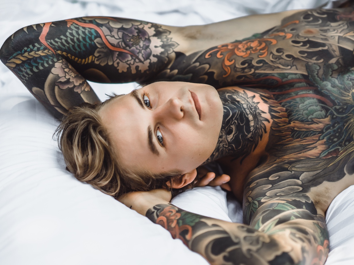 Красивый молодой человек с татуировками на теле лежит на кровати