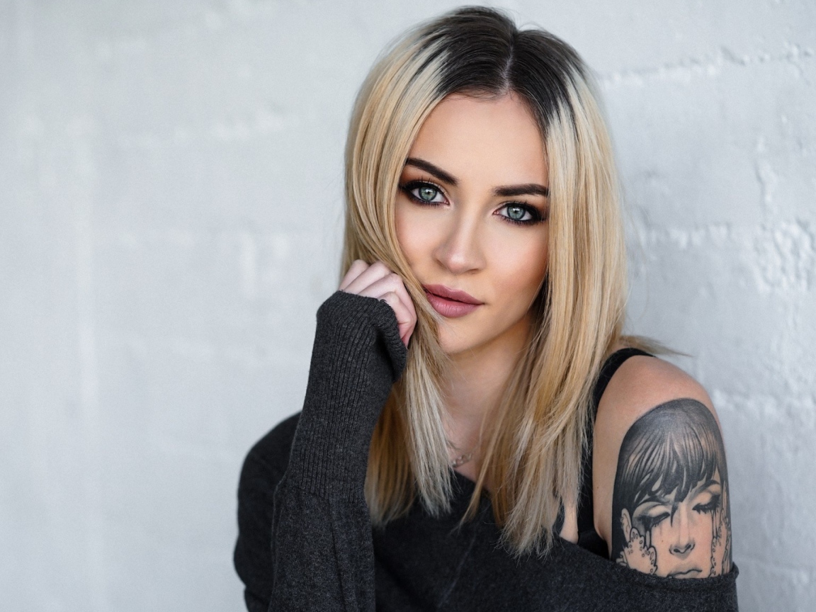 Стильная блондинка с татуировкой на плече на фоне стены