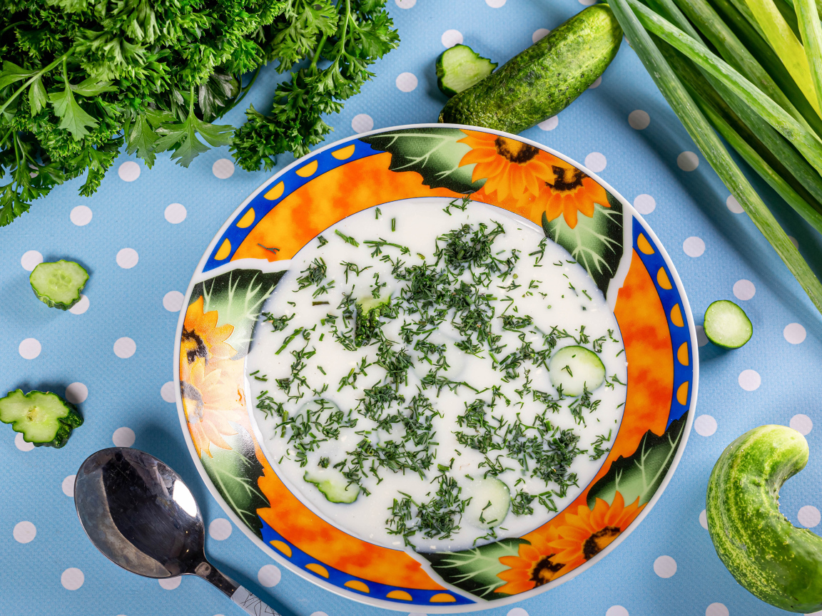 Окрошка в тарелке на столе с зеленью и огурцами