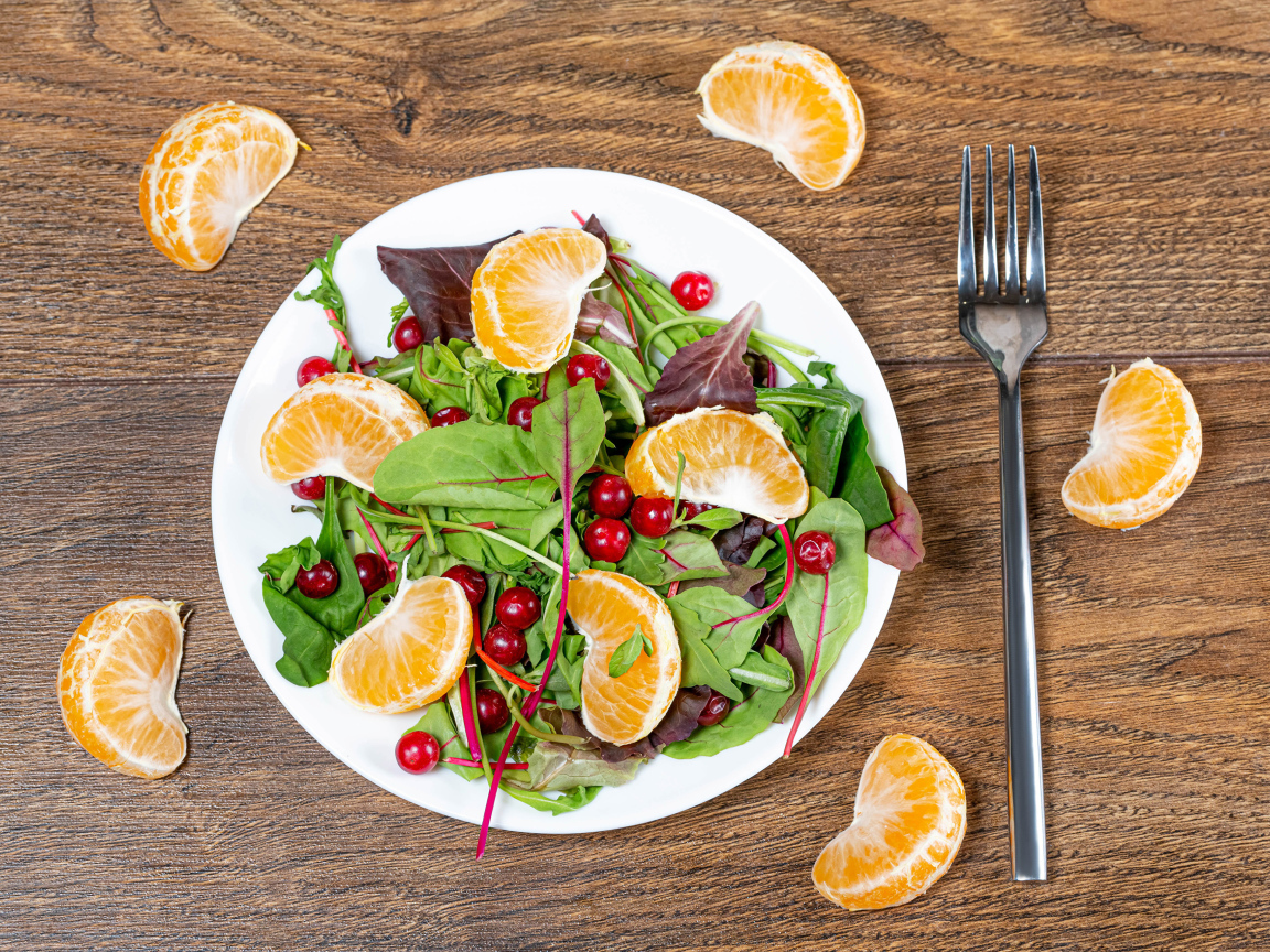 Салат из зелени и мандарина на белой тарелке на столе с вилкой