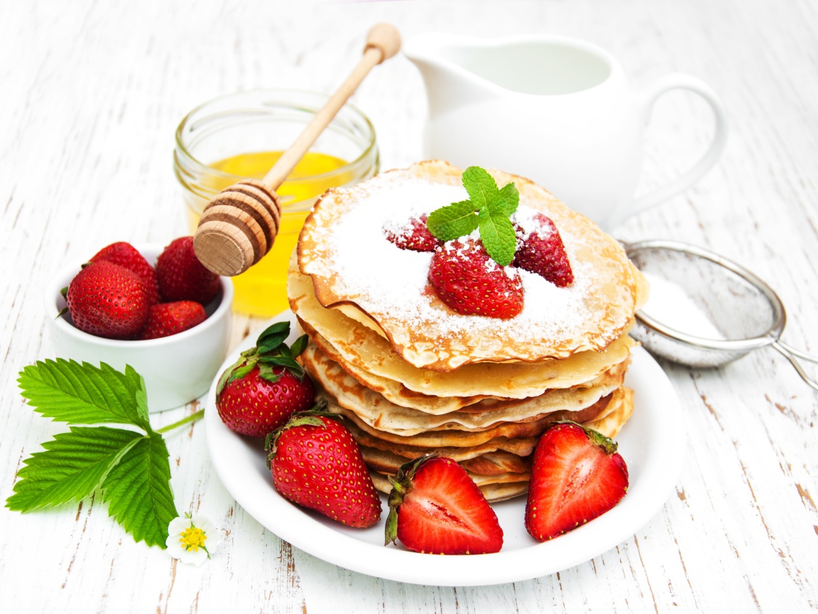 Тонкие блины с сахарной пудрой и ягодами клубники на столе с молоком и медом